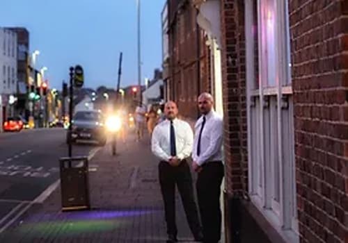 Two K1 door supervisors outside a venue on a town street at dusk